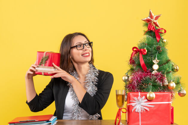 漂亮一位身着西装 戴着眼镜 面带微笑的商务女士手里拿着礼物 坐在一张桌子旁 桌子上放着一棵圣诞树人黑发微笑