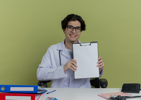 长袍面带微笑的年轻男医生 穿着医用长袍 戴着听诊器 戴着眼镜 坐在办公桌旁 手里拿着医疗工具 看上去很孤独医生微笑桌子