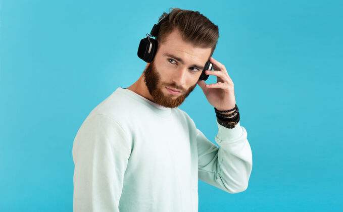 肖像时尚迷人的年轻胡须男子的肖像在无线耳机上听音乐现代风格自信的情绪在蓝色背景上孤立时尚帅气音频
