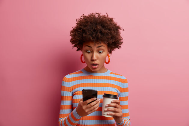肖像惊讶的卷发年轻女子盯着智能手机屏幕 在网上看到一些惊人的东西 阅读令人不安的侮辱性信息 喝外卖咖啡 对着粉色的墙壁摆姿势惊喜咖啡惊人