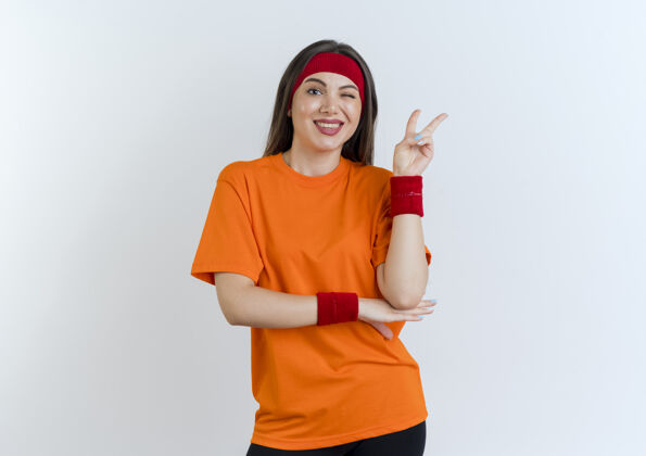 人微笑的年轻运动女性戴着头带和腕带把手放在手肘下眨眼做和平标志看起来孤立运动成年人腕带