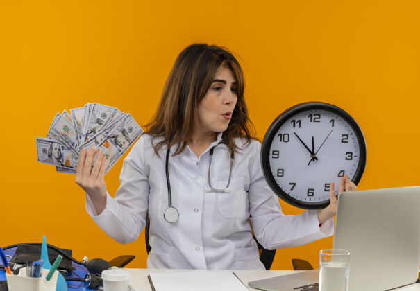 人印象深刻的中年女医生穿着医用长袍和听诊器坐在办公桌前 拿着医疗工具剪贴板和笔记本电脑拿着时钟和钱看时钟隔离时钟听诊器橙色