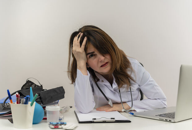 桌子疲惫的中年女医生穿着医用长袍和听诊器坐在办公桌旁 手里拿着医疗工具 剪贴板和笔记本电脑 手放在头上 看上去很孤立医疗工具坐着