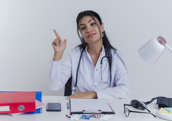 听诊器高兴的年轻女医生穿着医用长袍和听诊器坐在办公桌旁 手放在办公桌上抬起手指成年人女人手