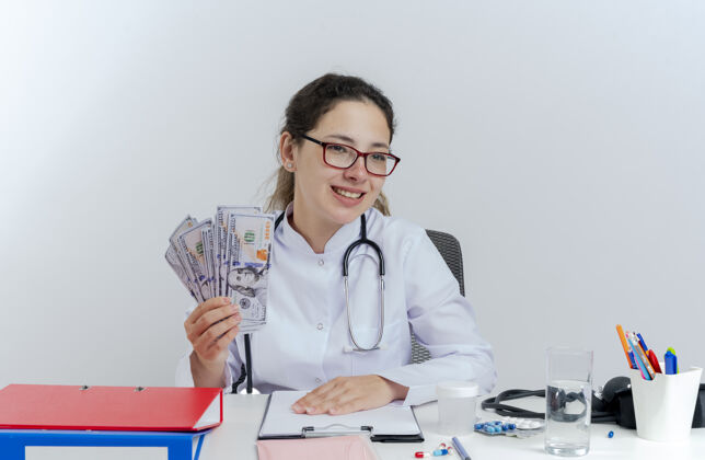 桌子面带微笑的年轻女医生穿着医用长袍 戴着听诊器和眼镜 坐在办公桌旁拿着医疗工具 手里拿着钱 一边孤立地看着听诊器长袍医生