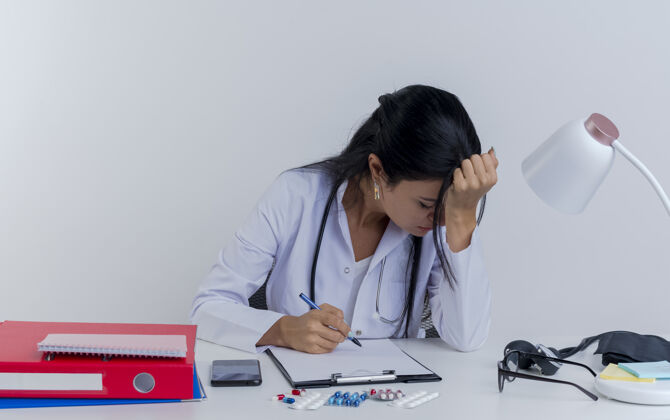 笔疲惫的年轻女医生穿着医用长袍 手持听诊器坐在办公桌旁 手持医疗工具 手放在头上 闭着眼睛 与世隔绝累了医生女人