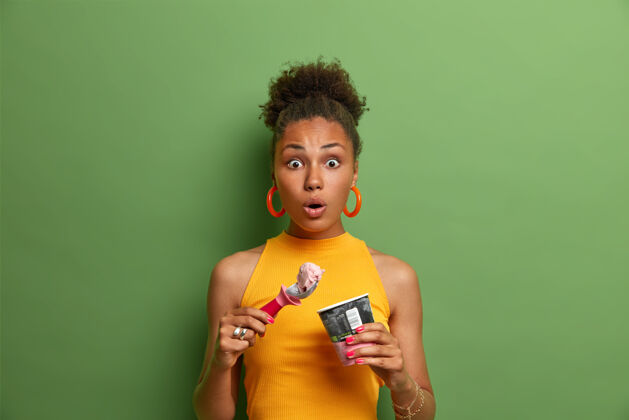 惊奇甜食和诱惑的概念无言惊讶的非裔美国妇女吃美味的冷甜点 享受草莓口味的冰淇淋 穿着黄色的夏装 绿色的墙壁印象姿势女士