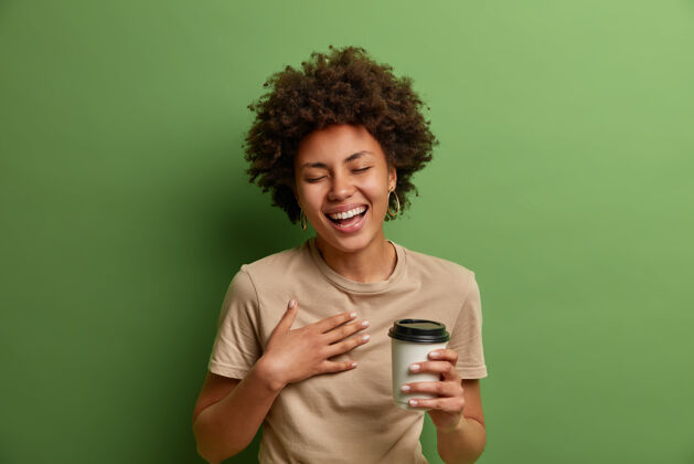 民族快乐的民族女孩的画像有乐趣和大声笑出来 举行一次性咖啡杯 微笑广泛和闭上眼睛 有愉快的交谈 隔离在绿色的墙壁生活方式的概念牙齿卷曲的