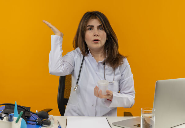 桌子困惑的中年女医生穿着医用长袍和听诊器坐在办公桌旁 拿着医用工具剪贴板和手提电脑 手里拿着医用烧杯 显示双手空空手工具烧杯