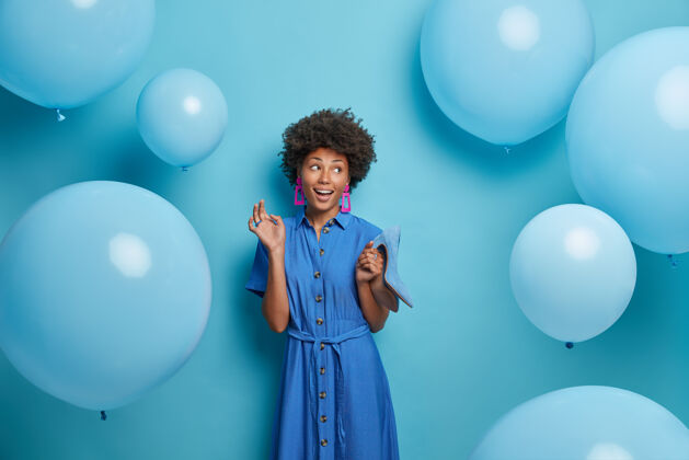 充气笑容可掬的美籍黑人妇女为生日聚会选择服装 高跟鞋上穿着蓝色的鞋子 高兴地向旁边看 在飞来飞去的充气气球旁摆姿势女人 衣服裙子时尚衣柜