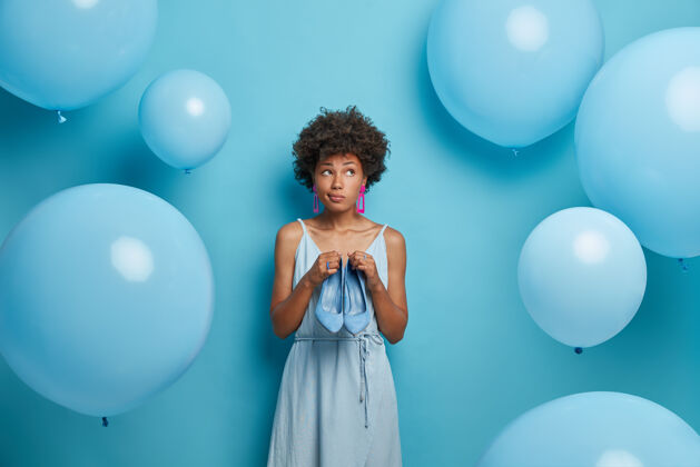 衣柜一个体贴的女性形象 穿着非洲头发的礼服参加派对 想穿什么更好 穿着蓝色的裙子 穿着高跟鞋 等待一些特别的东西 对着充气的气球摆姿势风格外观庆典