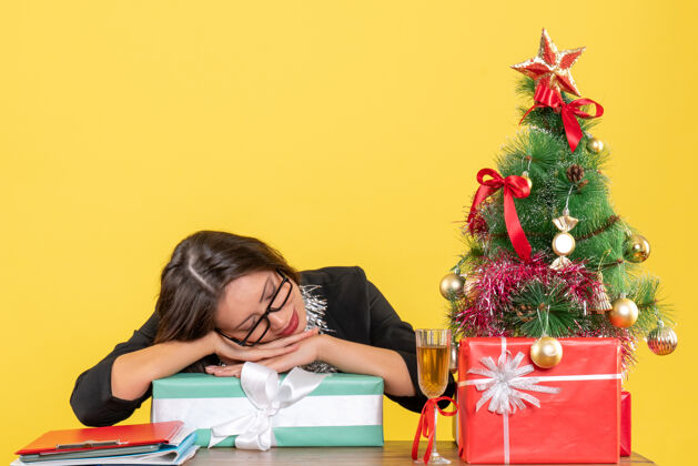 性感一位身着西装 戴着眼镜 面带微笑的商务女士睡在礼物上 坐在办公室的一张桌子旁 桌上放着一棵圣诞树礼物时尚快乐
