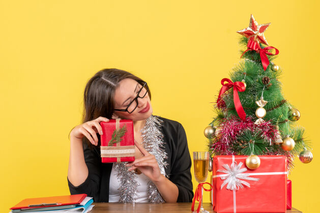 树一位穿着西装 戴着眼镜的梦幻商务女士手里拿着礼物 坐在一张桌子旁 桌子上放着一棵圣诞树快乐人眼镜