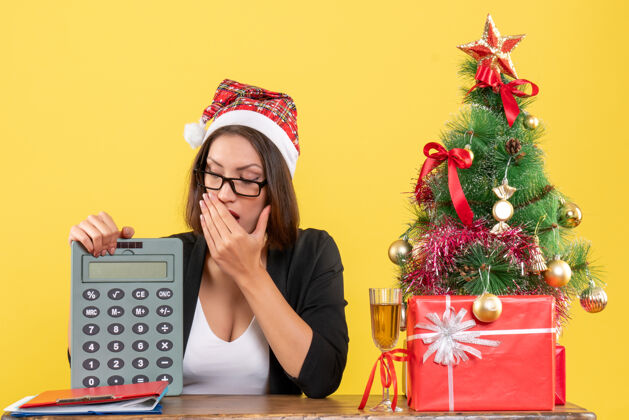 圣诞老人惊艳迷人的女士身着西装 戴着圣诞老人的帽子和眼镜 在黄色的办公室里展示计算器电话人计算器
