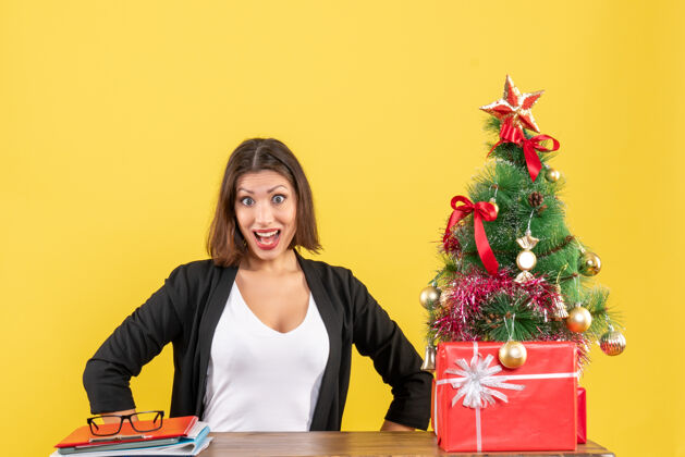 成人震惊的年轻女子穿着西装坐在一张桌子旁装饰圣诞树在黄色办公室黑发封面女郎性感