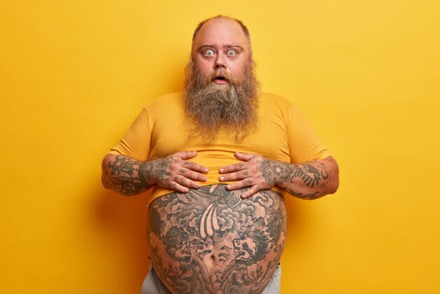 纹身惊诧的时髦男人手放在肚子上 t恤上有刺青 惊讶地发现自己的体重 长着浓密的胡须 靠着黄色的墙壁摆姿势男人露出了大肚子阳刚厚表情