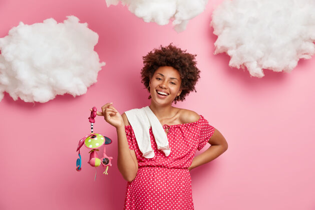 肖像美丽开朗的孕妇准备做母亲 大肚子 为未出生的孩子买玩具和衣服 享受快乐的期望 孤立在粉红色的墙上 上面有白云产妇微笑期望