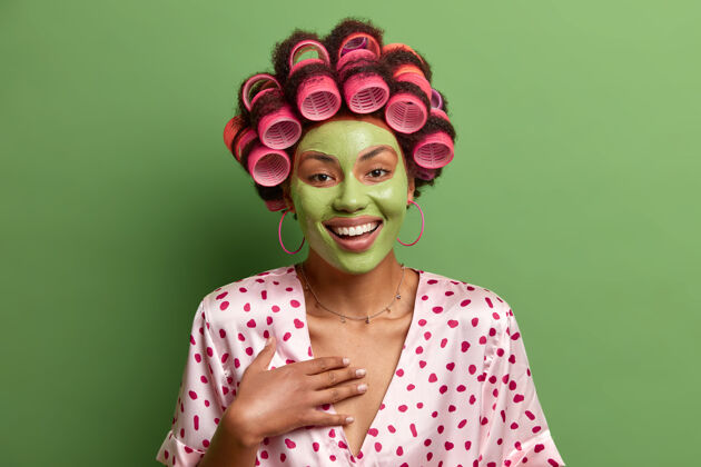 室内快乐无忧无虑的女人在家里享受美容的过程 为健康的皮肤穿绿色的面具 戴着卷发器 穿着丝绸长袍 听到有趣的东西 摆在室内长袍清洁牙齿
