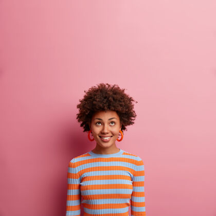 微笑欢快的民族女孩 卷发 聚精会神 心情很好 好奇地向上看 穿着休闲的条纹套头衫 孤零零地站在粉红色的墙上 空荡荡的非洲乐观发型