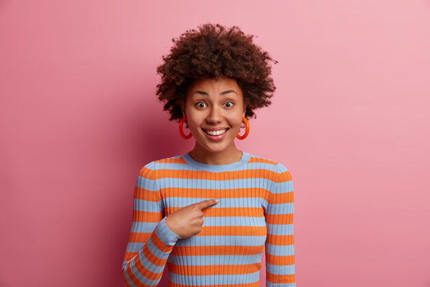 成人画像中开朗的卷发女孩积极微笑 被邀请参加 被人指着自己 被选中或提拔 穿着条纹毛衣 孤立在粉红色的墙上满意不确定谁