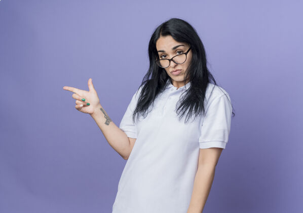 年轻自信的年轻黑发白种人女孩戴着眼镜指着隔离在紫色墙上的一侧女人壁板自信