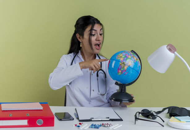 成人令人印象深刻的年轻女医生穿着医用长袍和听诊器坐在办公桌旁 手持医疗工具 看着并指着地球绿色指着医生