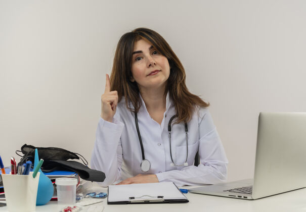 人令人印象深刻的中年女医生穿着医用长袍和听诊器坐在办公桌旁 拿着医疗工具 剪贴板和笔记本电脑 抬起手指 看上去与世隔绝听诊器剪贴板女士