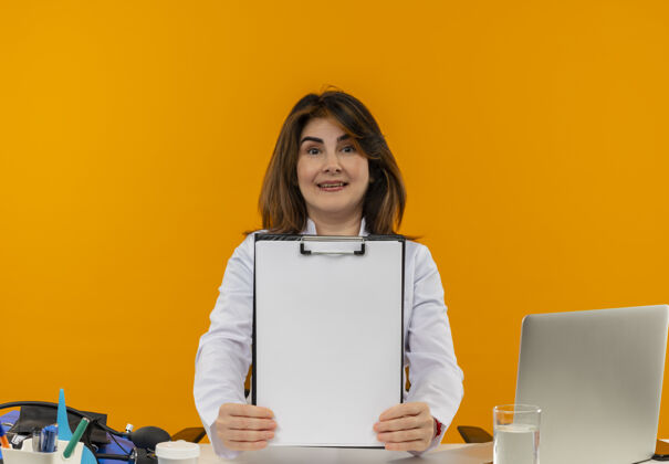 笔记本电脑微笑的中年女医生穿着医用长袍和听诊器坐在办公桌前 手里拿着医疗工具和笔记本电脑成人医疗表演