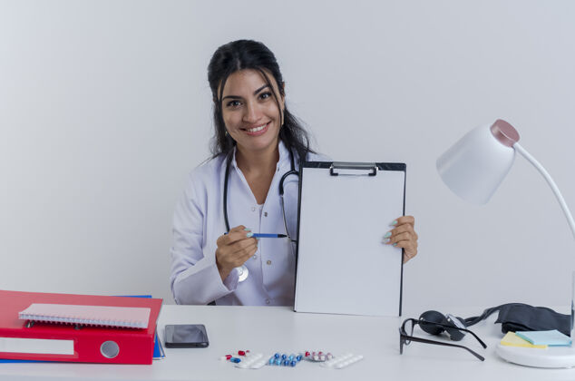 医生面带微笑的年轻女医生穿着医用长袍和听诊器坐在办公桌旁 手里拿着医疗工具 看着剪贴板 用钢笔指着它穿桌子工具