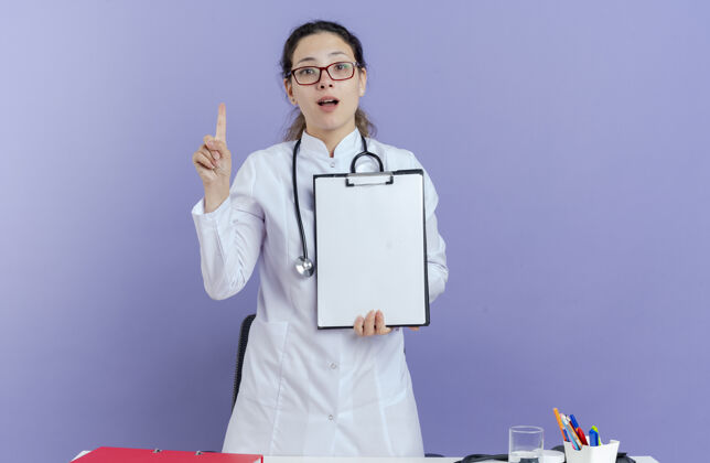 紫色年轻的女医生穿着医用长袍 戴着听诊器 戴着眼镜 站在桌子后面 手里拿着医疗工具 抬起手指 看着我女性医生女士