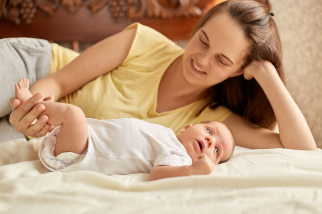 睡眠妈妈和宝宝躺在床上的白色毯子上 微笑着的妈妈穿着黄色的t恤 享受着和她刚出生的孩子在一起的时光 宝宝看着别处学习外面的东西新生儿儿子婴儿
