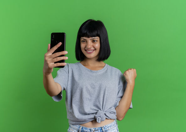 黑发微笑的年轻黑发白人女孩看着手机 保持拳头在绿色背景与复制空间隔离绿色电话年轻