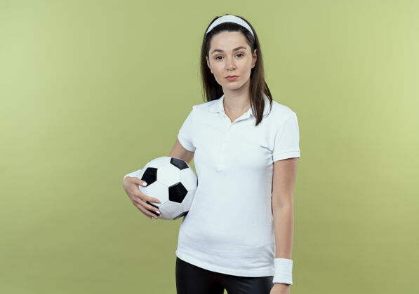 持球戴着头巾 拿着足球 表情严肃自信的年轻健身女士站在灯光背景下看着摄像机年轻表情站立