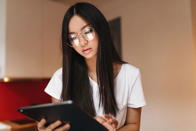 空气沉思的亚洲女人看着平板电脑的屏幕眼镜应用程序室内