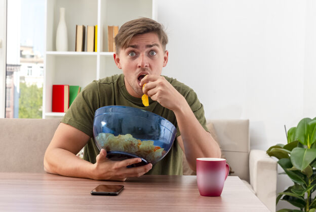 薯片震惊的年轻金发帅哥拿着杯子和电话坐在桌子旁 手里拿着一碗薯片 在客厅里吃着坐着拿着客厅