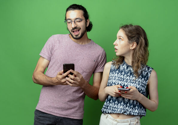 惊喜一对年轻夫妇拿着智能手机 看着绿色背景下的男友 女人既惊讶又困惑困惑男朋友手机