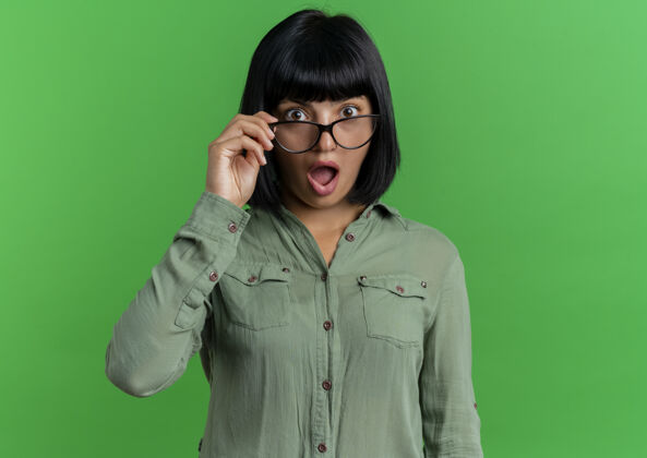 女孩兴奋的年轻黑发白人女孩手持光学眼镜 看着隔离在绿色背景和复印空间的相机复制年轻眼镜