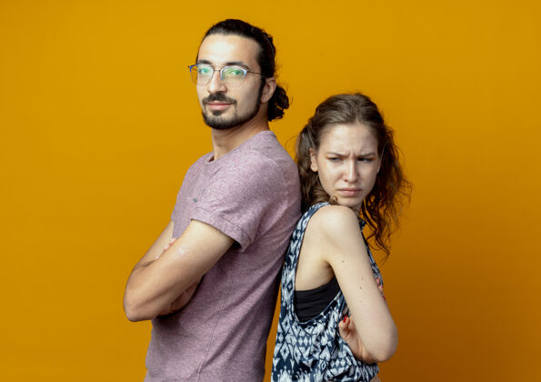 情侣一对年轻的夫妇看着摄像机背靠背地站在橙色背景上 双臂交叉站立镜头年轻人