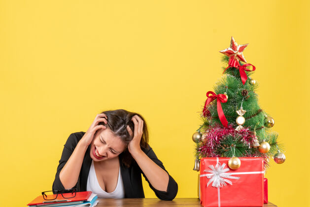 靠近愤怒 疲惫 紧张的年轻女子坐在黄色办公室装饰的圣诞树旁的桌子旁女士漂亮时尚