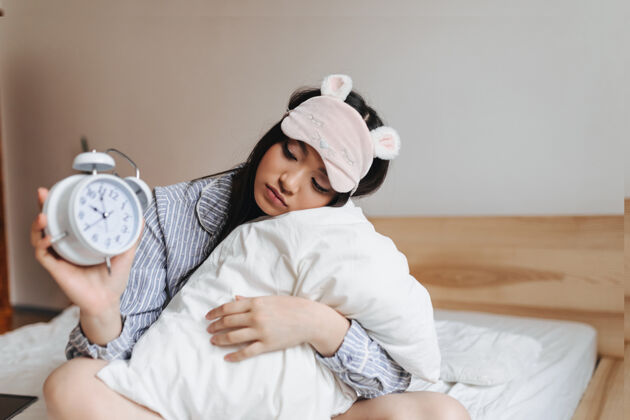 可爱戴着粉色睡眠面具的女人抱着枕头 悲伤地看着闹钟女人睡眠面具面具