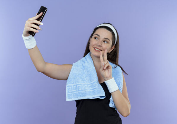 头带戴着头巾脖子上围着毛巾的年轻健身女士看着智能手机的屏幕自拍显示胜利歌唱微笑着站在蓝色背景上屏幕脖子站