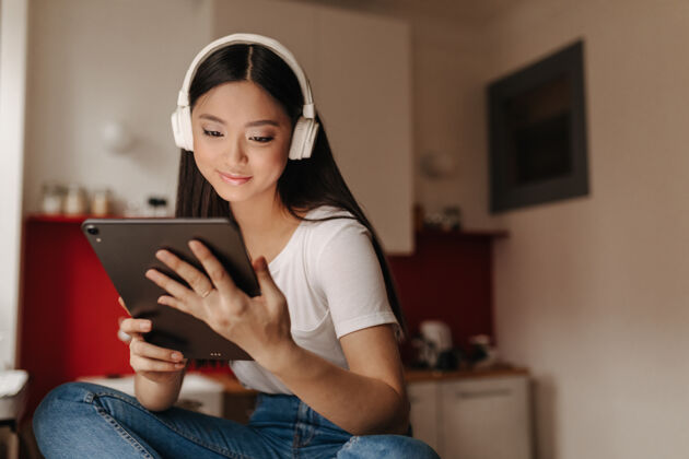 桌子穿着牛仔裤和白色t恤的年轻女子看着平板电脑 戴着耳机听音乐歌曲房子年轻