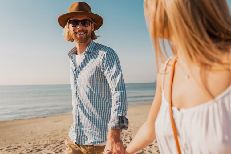 眼镜年轻迷人的微笑着戴着帽子的快乐男人和穿着白裙子的金发女人一起在沙滩上奔跑旅行旅行沙滩