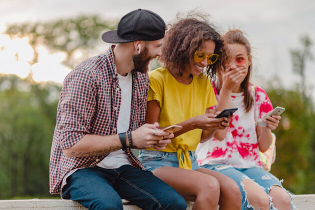 学生快乐的年轻朋友们坐在公园里用智能手机微笑 男人和女人玩得开心年轻青春手机