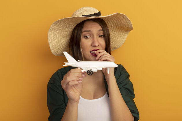 姿势焦虑的漂亮女人戴着沙滩帽咬着钉子拿着模型飞机看着隔离在橙色墙上帽子女人站