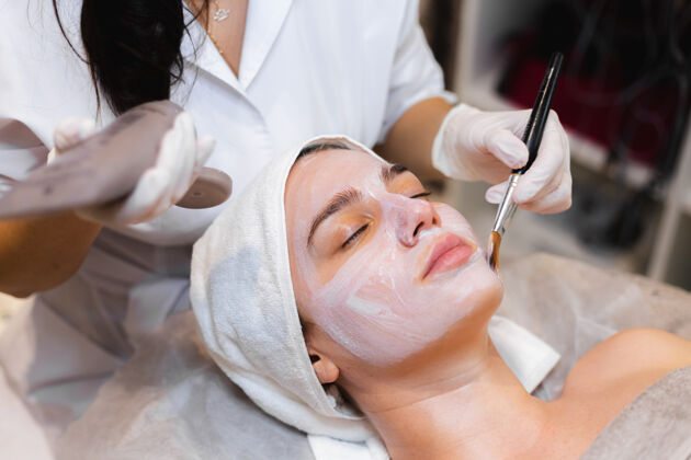 女士美容师用刷子将白色保湿面膜涂在spa美容院一位年轻女客户的脸上女人健康面部