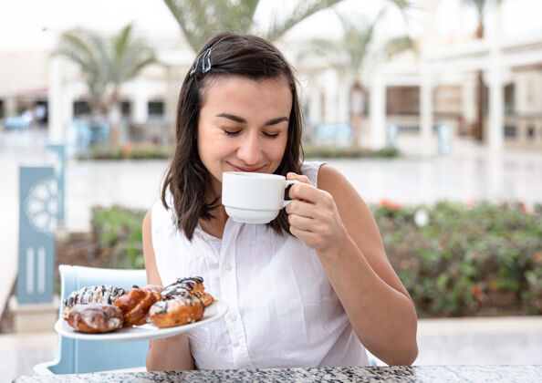 露台快乐的年轻女子在户外露台上享受着早晨的咖啡和甜甜圈咖啡馆放松美味