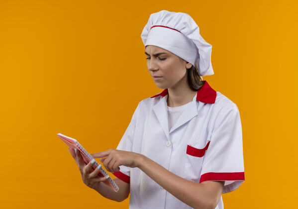 厨师穿着厨师制服的年轻白种人厨师女孩拿着笔记本 看着孤立在橙色背景上的有复印空间的笔记本拿着笔记本烹饪