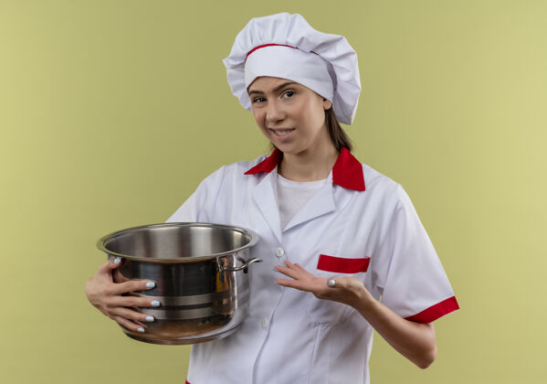 锅穿着厨师制服的年轻白种人厨师女孩拿着并指着绿色背景上有复制空间的孤立锅烹饪恼火制服