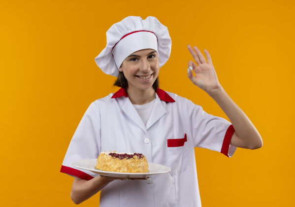 厨师身着厨师制服 面带微笑的白人年轻厨师女孩拿着盘子上的蛋糕 在橙色背景上做手势“ok”手势 并留有复印空间盘子手势蛋糕